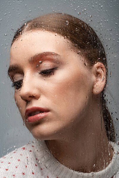 beautiful lonely woman on grey, beyond wet glass with drops 