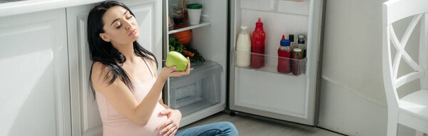 panoramic shot of tired pregnant woman looking at apple while sitting on floor in kitchen near opened fridge