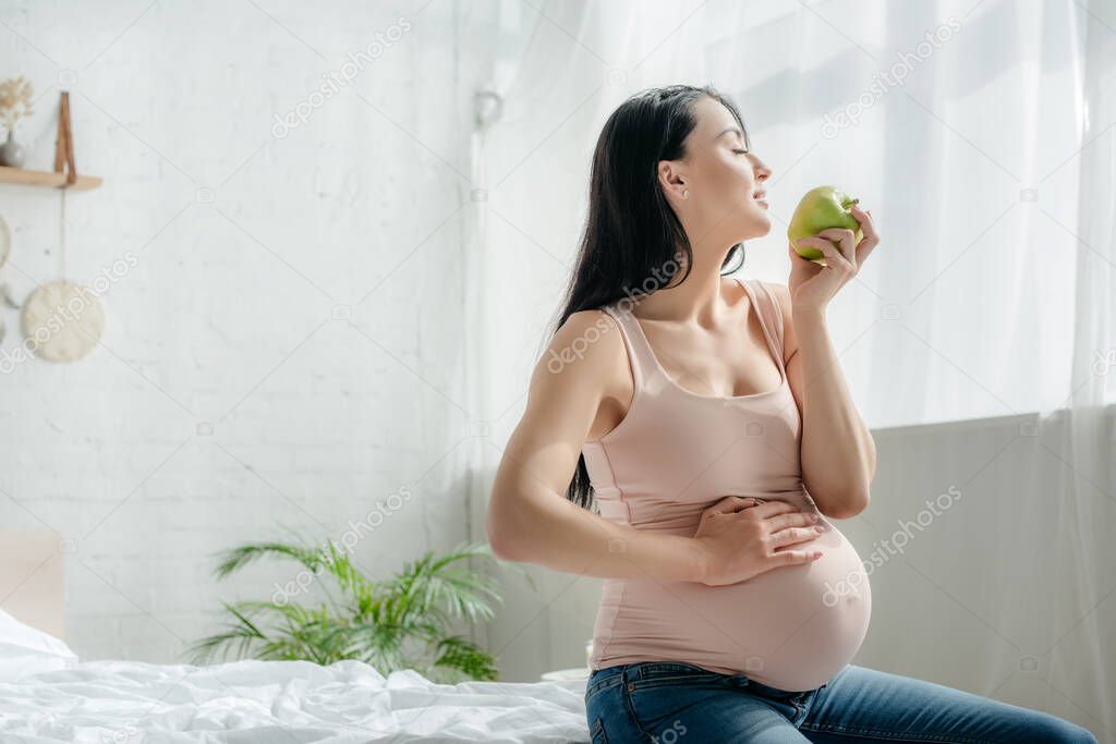beautiful pregnant woman holding apple and touching belly while sitting on bed 