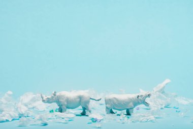 White toys of hippopotamus and rhinoceros with plastic garbage on blue background, animal welfare concept clipart