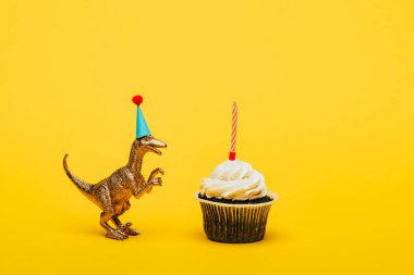 Parti şapkalı oyuncak dinozor ve sarı arka planda mumlu kek.