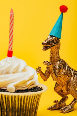 Oyuncak dinozorun parti şapkasındaki seçici odağı sarı arka planda mumu olan kek yanında.