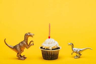 Toy dinosaurs beside cupcake with candle on yellow background clipart