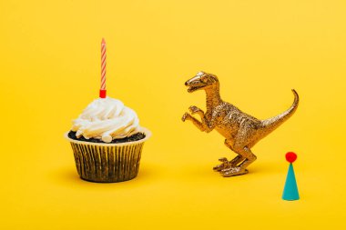 Parti şapkasının yanında oyuncak dinozor ve sarı arka planda mumu olan kek.