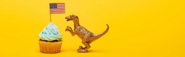 Oyuncak dinozorun panoramik görüntüsü kapkekin yanında sarı arka planda Amerikan bayrağı var.