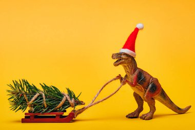 Sarı arka planda çam ağacıyla Noel Baba şapkalı oyuncak dinozor.