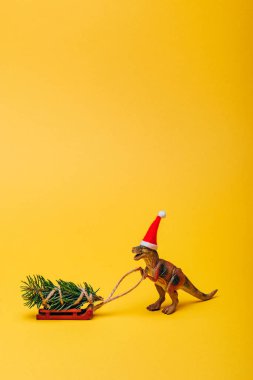 Noel Baba şapkalı oyuncak dinozor. Sarı arka planda kızak üzerinde köknar var.