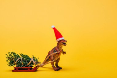 Noel Baba şapkalı oyuncak dinozor. Sarı arka planda kızak üzerinde köknar var.
