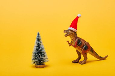 Noel Baba şapkalı oyuncak dinozor sarı arka plandaki Noel ağacının yanında.