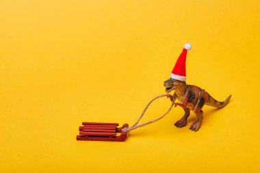 Toy dinosaur in santa hat with sleigh on yellow background clipart