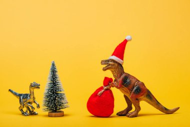 Sarı arka planda çam ağacının yanında Noel Baba şapkası ve çuvalı olan oyuncak dinozorlar.
