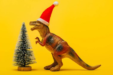 Noel Baba şapkalı oyuncak dinozor sarı arka plandaki Noel ağacının yanında.