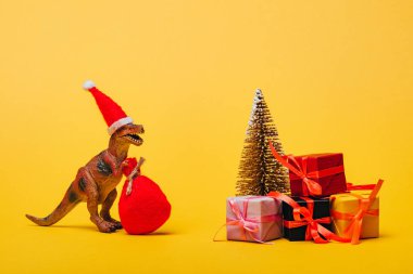 Noel Baba şapkalı oyuncak dinozor ve çam ağacının yanında sarı arka planda hediyeler.