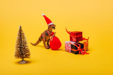 Noel Baba şapkalı oyuncak dinozor ve çam ağacının yanında sarı arka planda hediyeler.