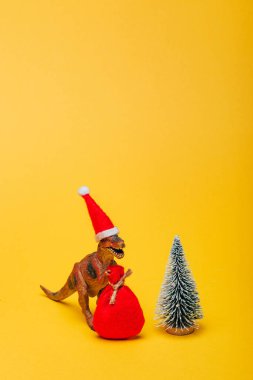 Noel Baba şapkalı oyuncak dinozor. Sarı arka planda Noel ağacı yanında çuval var.