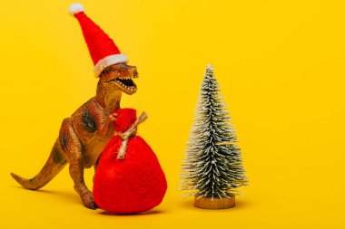 Noel Baba şapkalı oyuncak dinozor. Sarı arka planda Noel ağacı yanında çuval var.