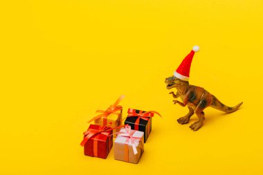 Noel Baba şapkalı oyuncak dinozor sarı arka planda hediyelerin yanında