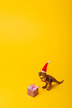 Sarı arka planda hediyeli Noel Baba şapkalı oyuncak dinozor.
