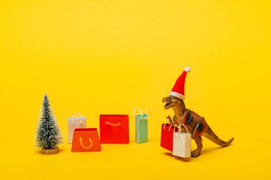 Noel Baba şapkalı oyuncak dinozor alışveriş torbaları ve sarı arka planda noel ağacı.