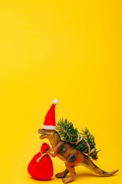 Noel Baba şapkalı oyuncak dinozor ve sarı arka planda köknar.