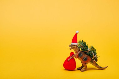 Noel Baba şapkalı oyuncak dinozor ve sarı arka planda köknar.