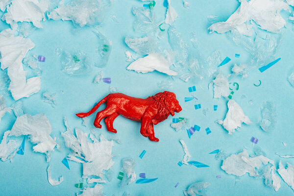 Top view of red toy lion with plastic garbage on blue background, environmental pollution concept