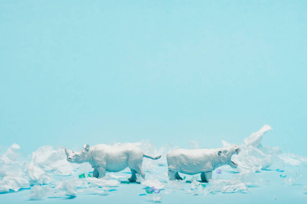 White toys of hippopotamus and rhinoceros with plastic garbage on blue background, animal welfare concept