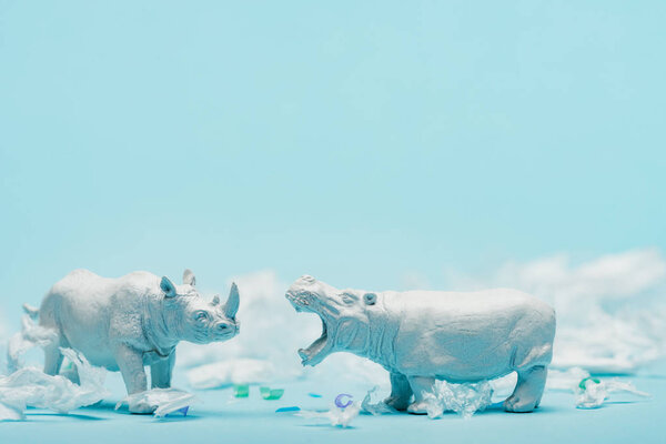 White hippopotamus and rhinoceros toys with plastic garbage on blue background, animal welfare concept