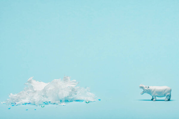 White hippopotamus toy and plastic garbage on blue background, animal welfare concept