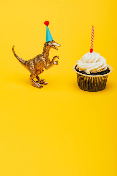 Toy dinosaur in party cap beside cupcake with candle on yellow background
