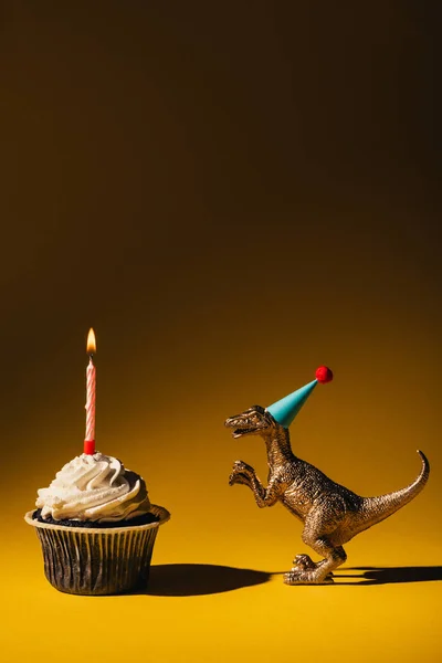 Zabawka Dinozaur Party Cap Obok Babeczki Płonącą Świecą Brązowym Tle — Zdjęcie stockowe