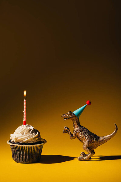 Toy dinosaur in party cap beside cupcake with burning candle on brown background