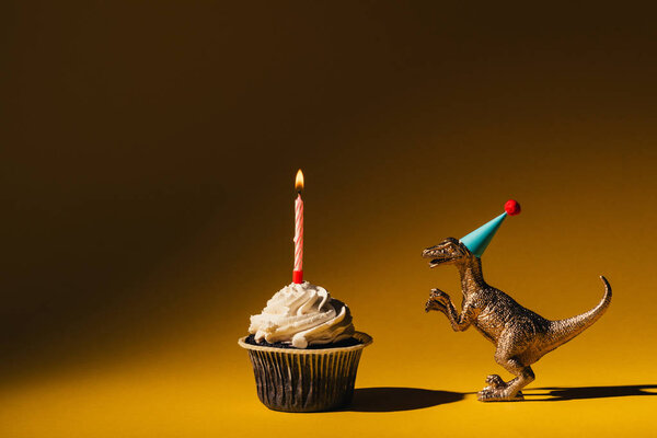 Игрушечный динозавр в кепке для вечеринок рядом с кексом с горящей свечой на коричневом фоне
