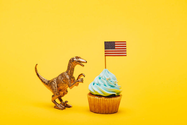 Toy dinosaur beside cupcake with american flag on yellow background