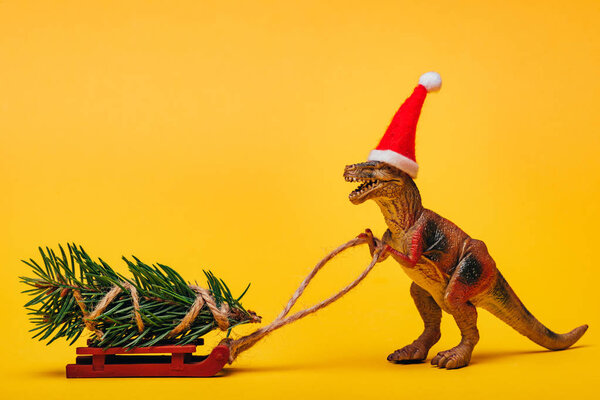 Toy dinosaur in santa hat with pine on sleigh on yellow background