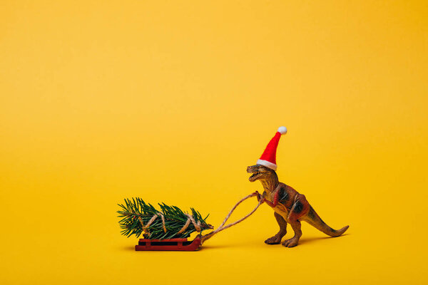 Toy dinosaur in santa hat with pine on sleigh on yellow background