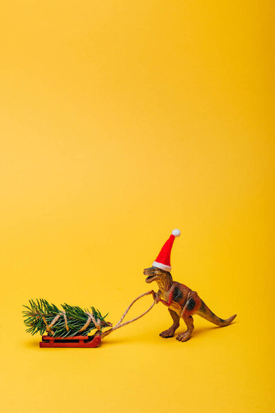 Toy dinosaur in santa hat with fir on sleigh on yellow background