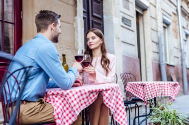 Caddedeki kafede oturup kırmızı şarap bardaklarını tokuştururken erkek arkadaşına bakan çekici genç bir kadın.