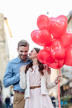 smiling man embracing happy girlfriend holding red heart-shaped balloons on street clipart