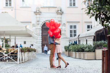Mutlu genç çift şehir meydanında kırmızı kalp şeklinde balonların arkasına saklanırken kucaklaşıyorlar.