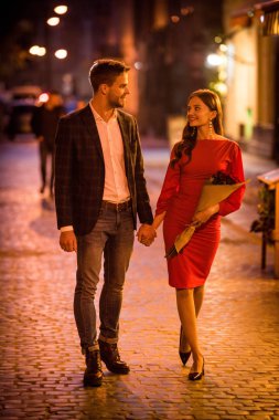 Mutlu, zarif bir çift el ele tutuşmuş ve gece sokakta yürürken birbirlerine bakıyorlar.