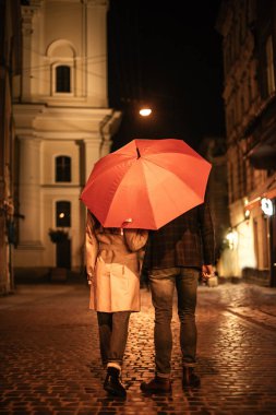 Sonbahar kıyafetli bir çiftin akşam caddesi boyunca şemsiye altında yürüdüğünü görüyorum.