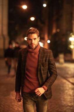 Sonbahar kıyafetli yakışıklı, kendine güvenen bir adam akşam sokakta yürürken kameraya bakıyor.