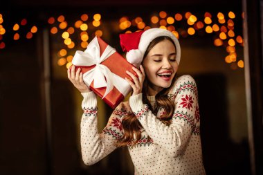 Sıcak süveter ve Noel Baba şapkalı, kapalı gözlerle hediye kutusunu tutan genç bir kadın.