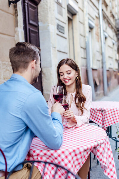 happy young woman clinking glasses of red wine with boyfriend while sitting in street cafe
