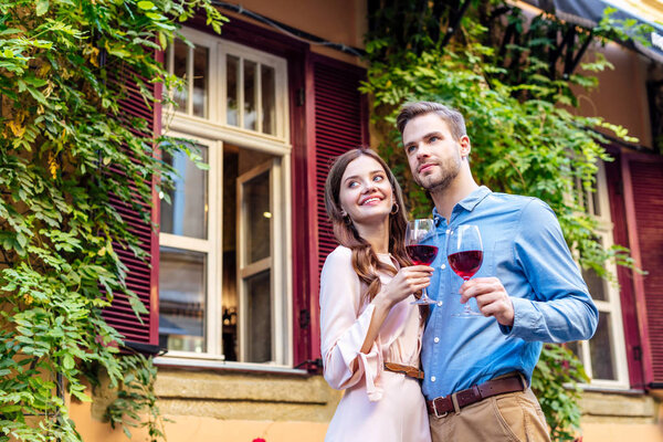 happy couple holding glasses of red wine while standing near house covered with green ivy and looking away