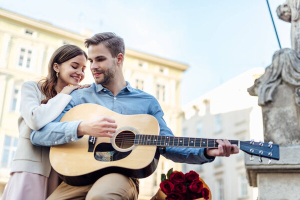 happy girl leaning on shoulder of boyfriend playing acoustic guitar on street