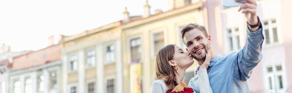 panoramic shot of young woman kissing boyfriend taking selfie on smartphone