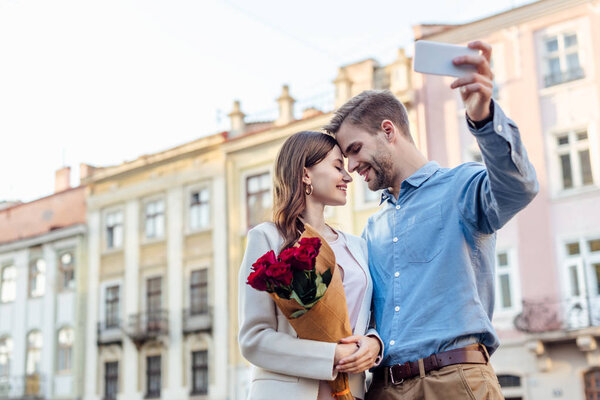 happy young woman holding bouquet of roses while smiling boyfriend taking selfie on smartphone