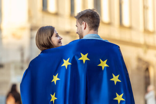 молодая пара туристов, завернутая в флаг Европейского союза, смотрящих друг на друга на улице
